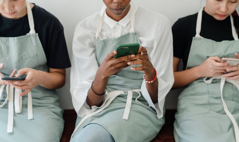 Three young café workers in aprons sit side by side, each engrossed in their smartphones during a break, creating a moment of relaxation and connectivity.