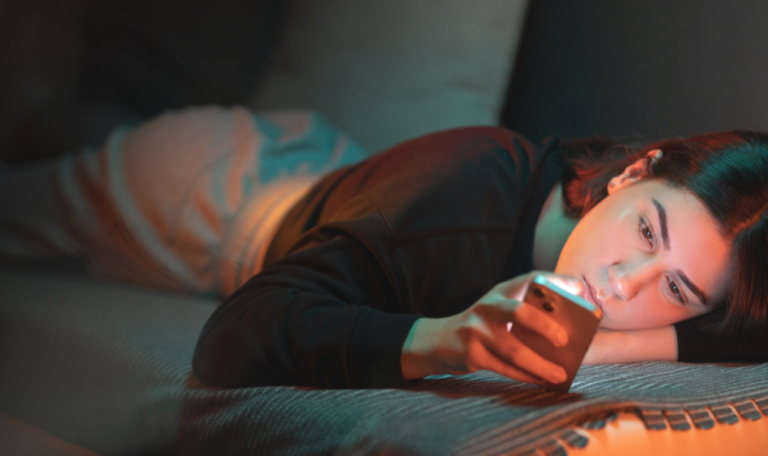 A girl lies on a couch, gazing at their smartphone in dim light, suggesting relaxation or late-night screen use.