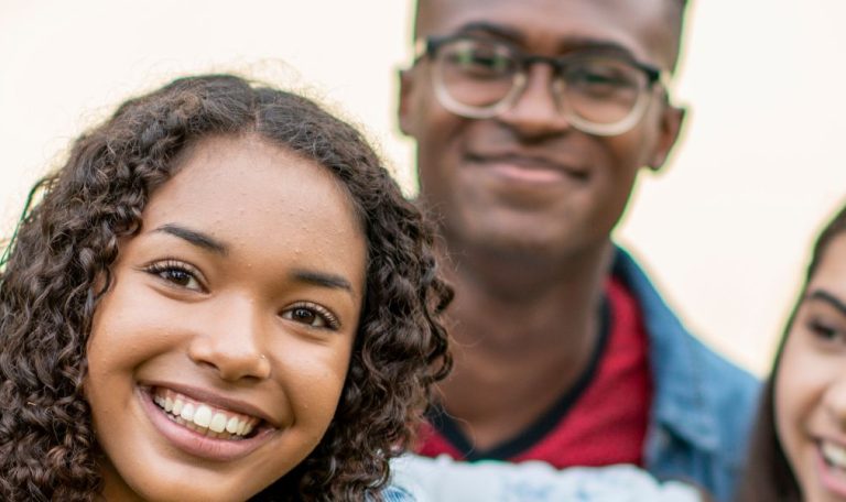 Three smiling teenagers standing close outdoors. The girl in front is beaming, with two friends behind her, sharing a joyful moment.