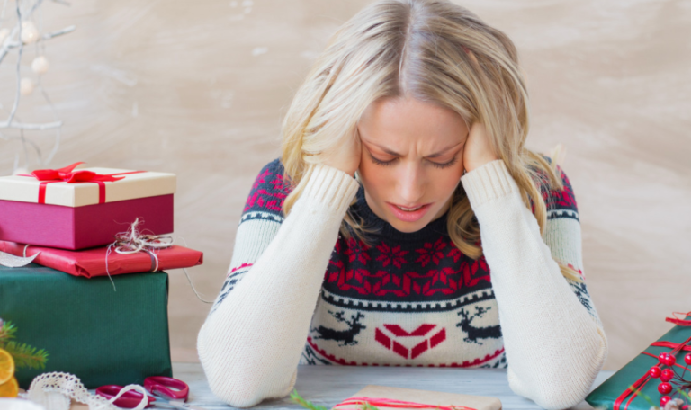 A woman in a festive sweater holds her head in frustration while sitting at a table filled with holiday gifts and decorations.