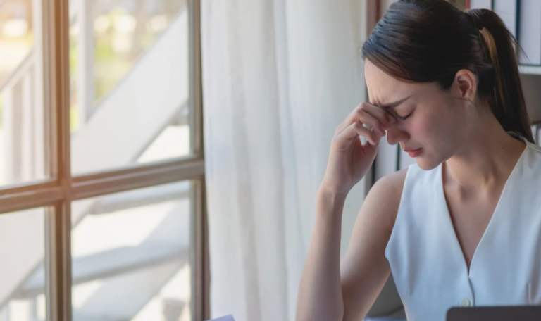 A woman sits at a desk with her hand on her forehead, overwhelmed by financial reports, charts, and stacks of documents."