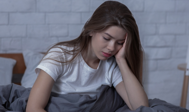 A young woman, sitting on a bed under grey blankets, looks tired and sad as she rests her head in her hand.