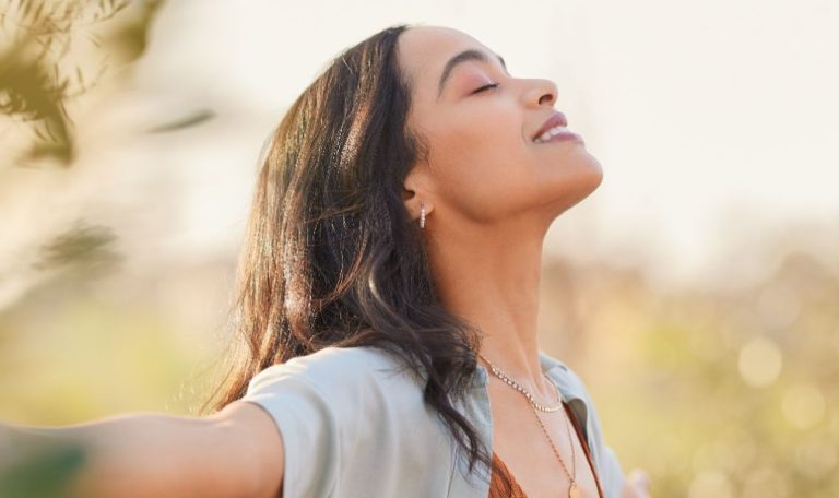 A woman stands outdoors, arms outstretched, enjoying the fresh air and sunlight with a peaceful, content expression on her face.