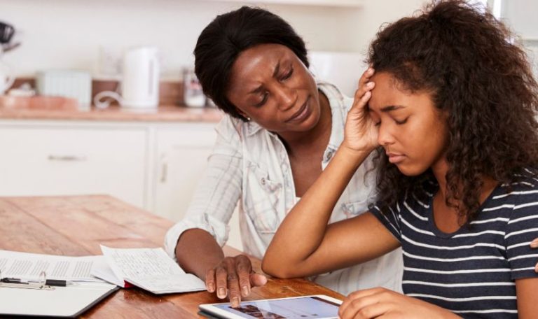 A mother sits beside her teenage daughter, offering comfort and support as the teen looks stressed and anxious while using a tablet at the table.
