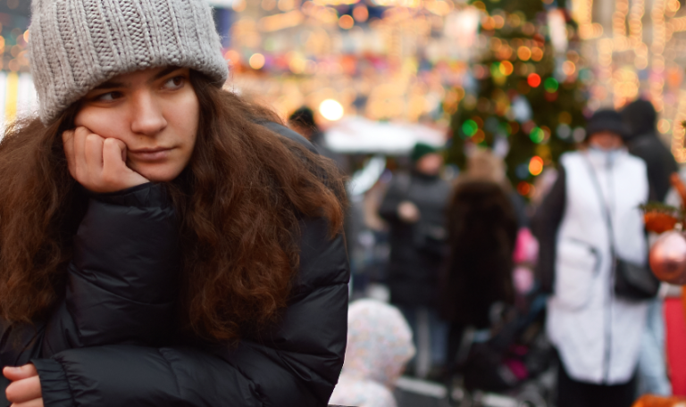 Woman in a knitted beanie, leaning on a table, looking bored amidst a lively Christmas market decorated with bright lights.