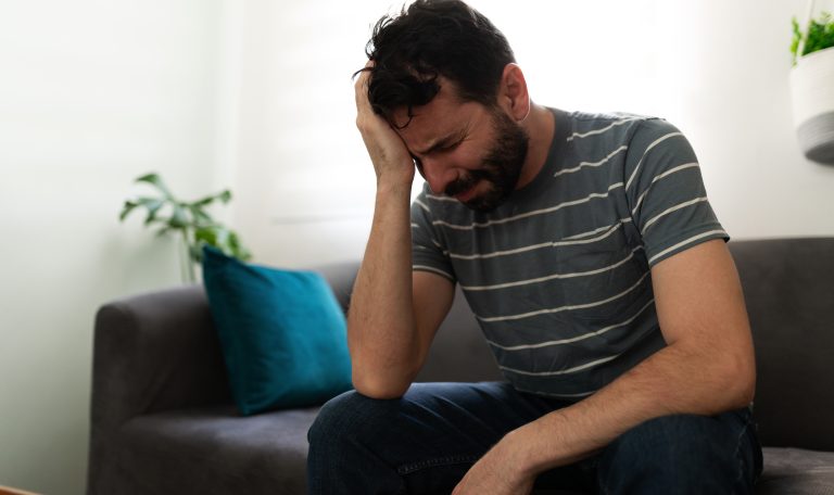 A man sitting on a couch holding his head in distress, representing emotional pain and men’s mental health challenges during Movember.